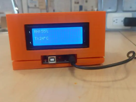 A small orange Arduino air quality monitoring device sits on a table, displaying “RH: 55%” and “T: 24°C” on its blue LCD screen, with a USB cable connected for power or data transfer.