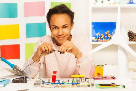 A young Black girl smiling while connecting jumper wires on an electronics circuit project kit at a desk with a white model wind turbine