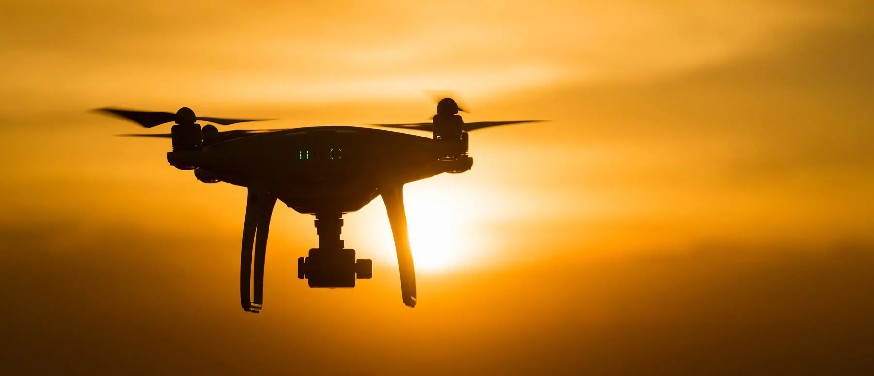 A silhouette of a quadcopter drone hovering in flight against a bright orange sunset sky