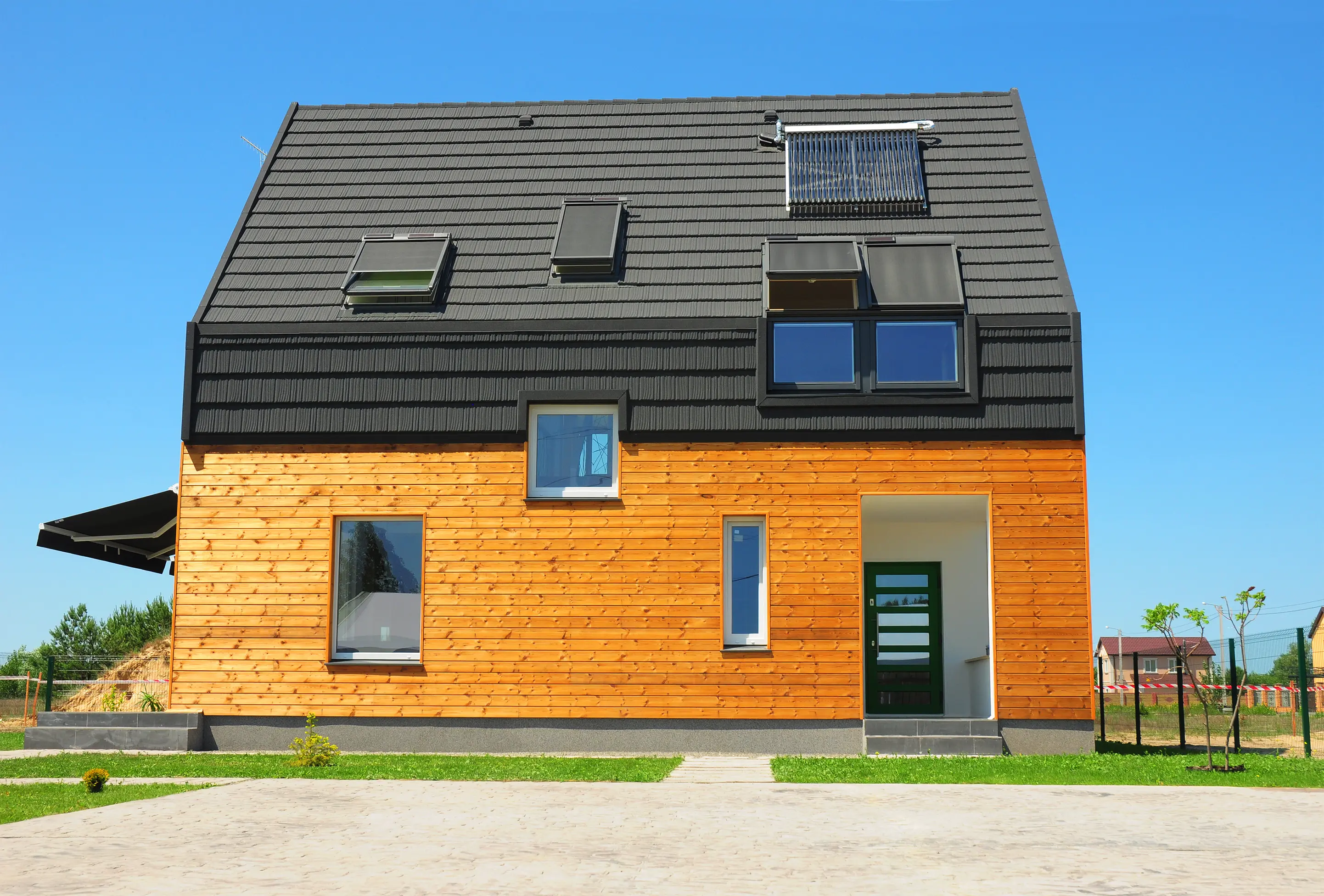 A modern two-story house featuring a light-colored wooden facade on the lower level and a dark grey gabled roof with integrated windows and solar collectors under a clear blue sky