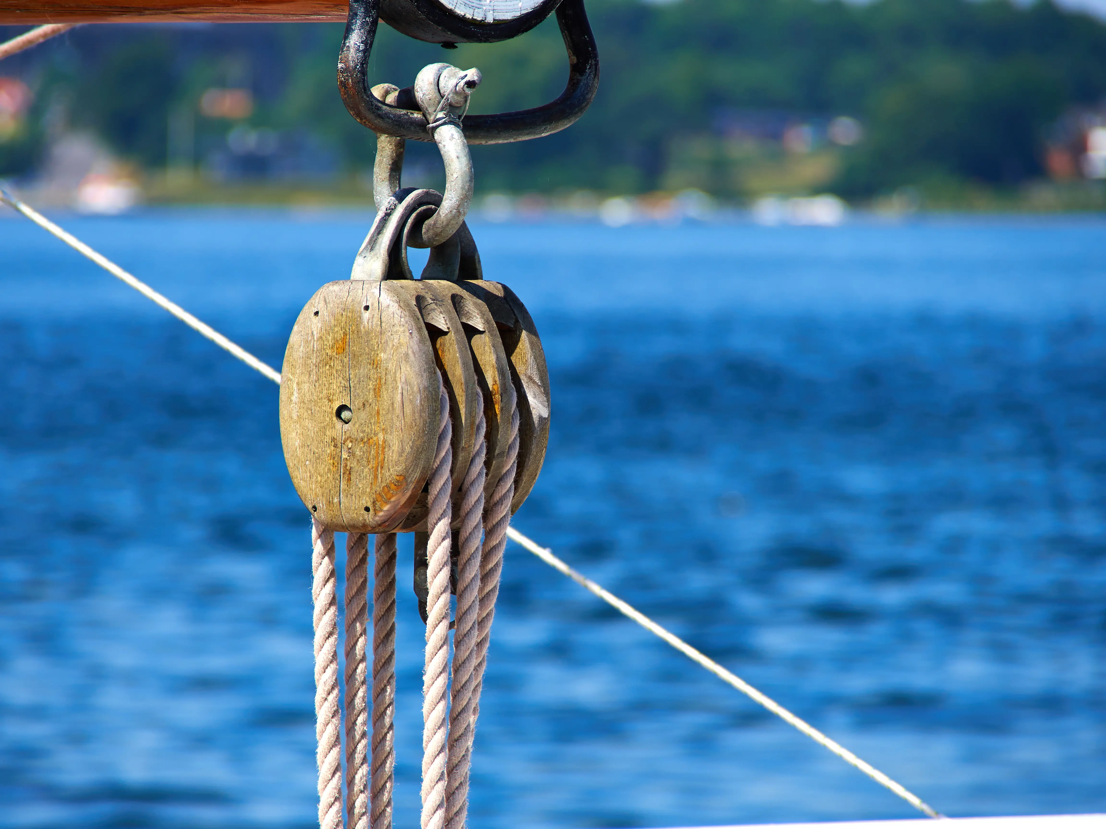A close-up view of a classic wooden triple-sheave block and tackle pulley system rigged with ropes on a boat with blue water blurred in the background