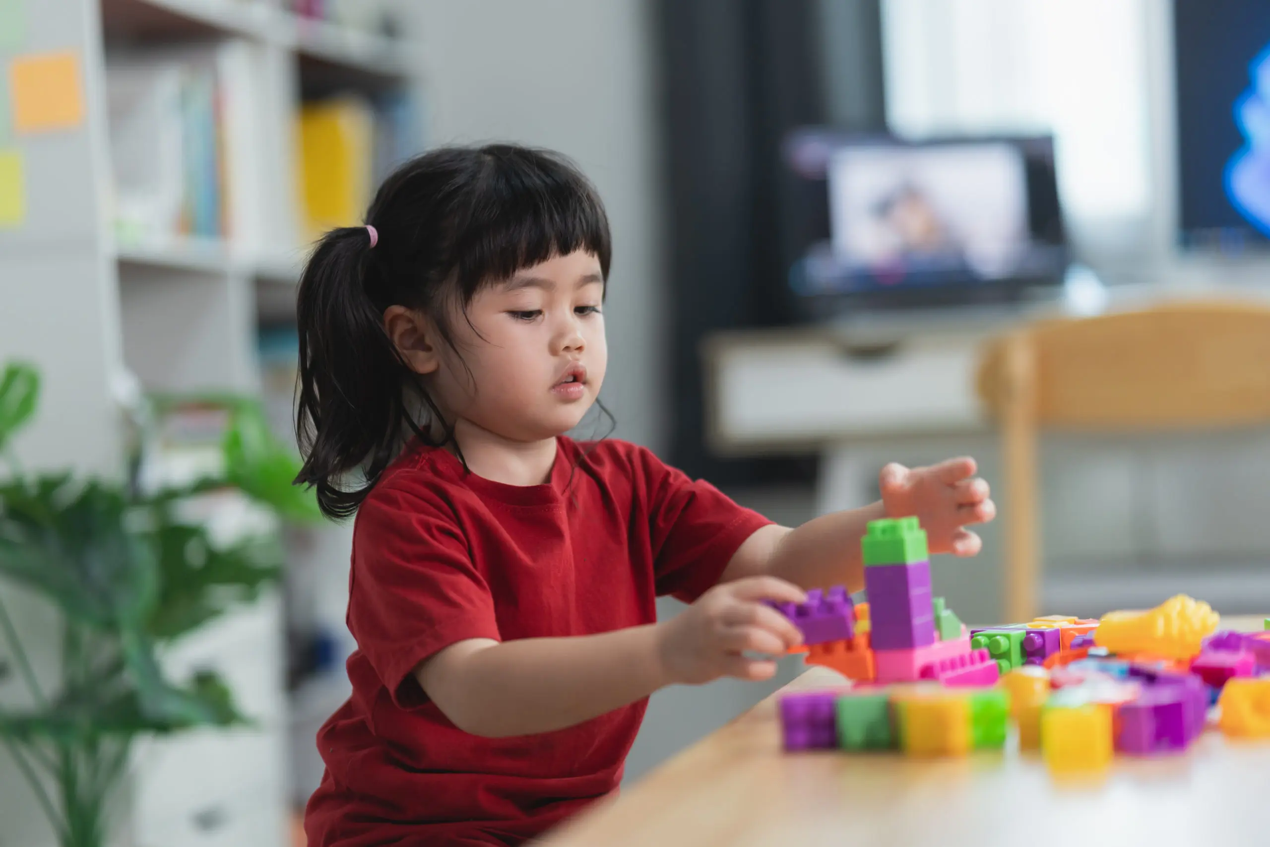 A young girl with pigtails in a red shirt playing with colorful interlocking plastic building blocks at a wooden table