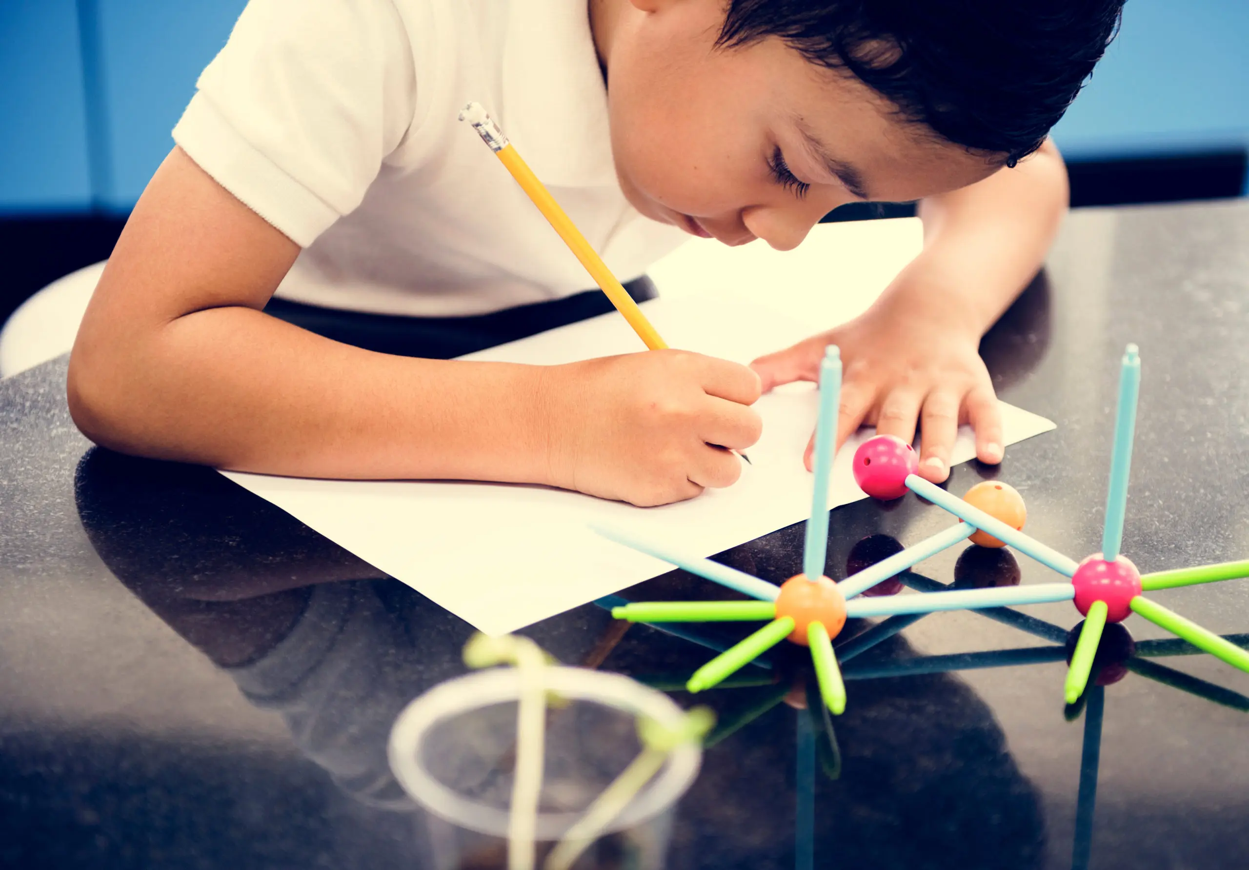 A young boy leans over a dark table writing on a white sheet of paper with a yellow pencil next to plastic molecular model toys