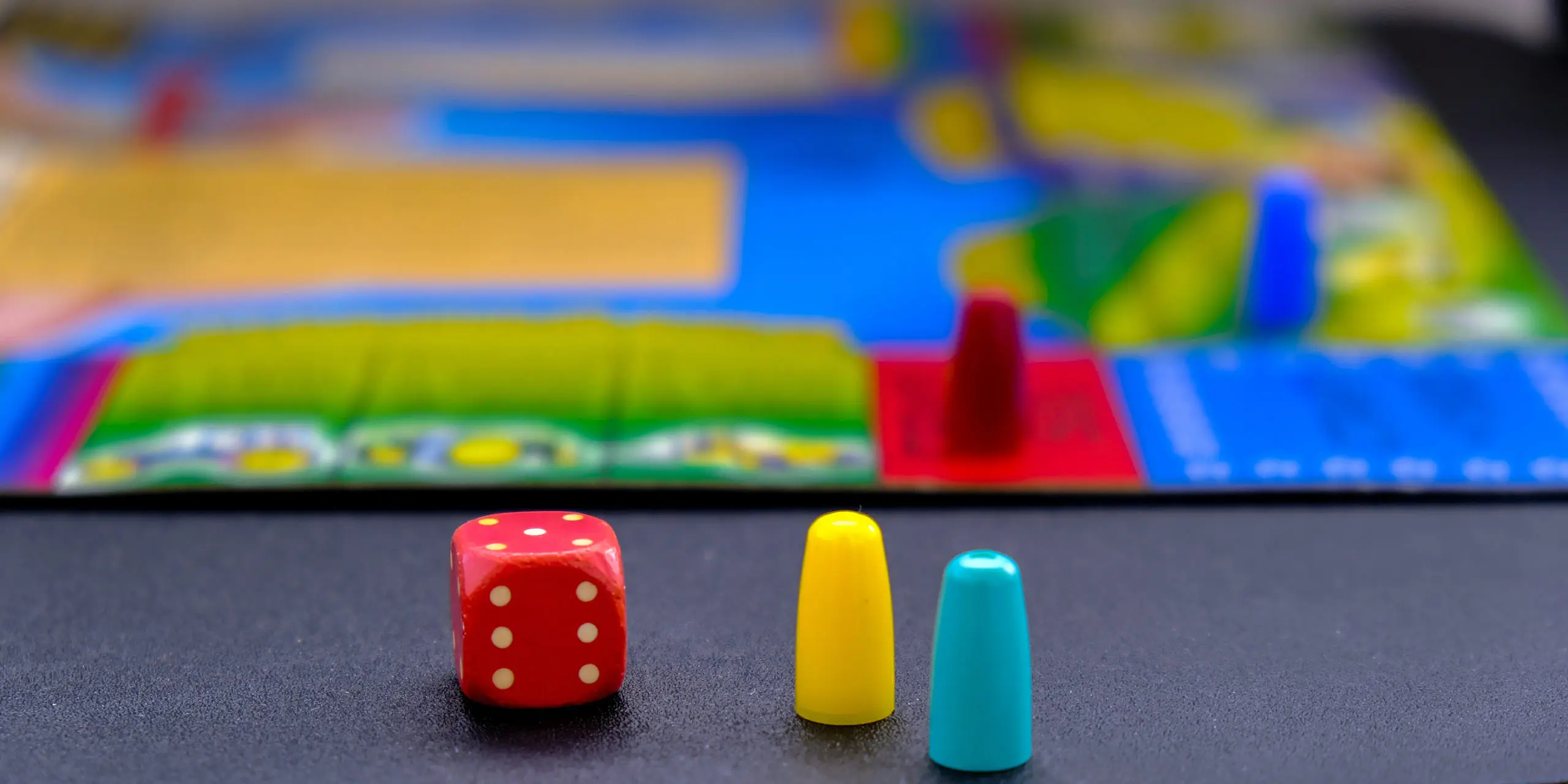 A red six-sided die showing six dots sits next to a yellow and a light blue game pawn on a dark surface in front of a blurred colorful children's board game