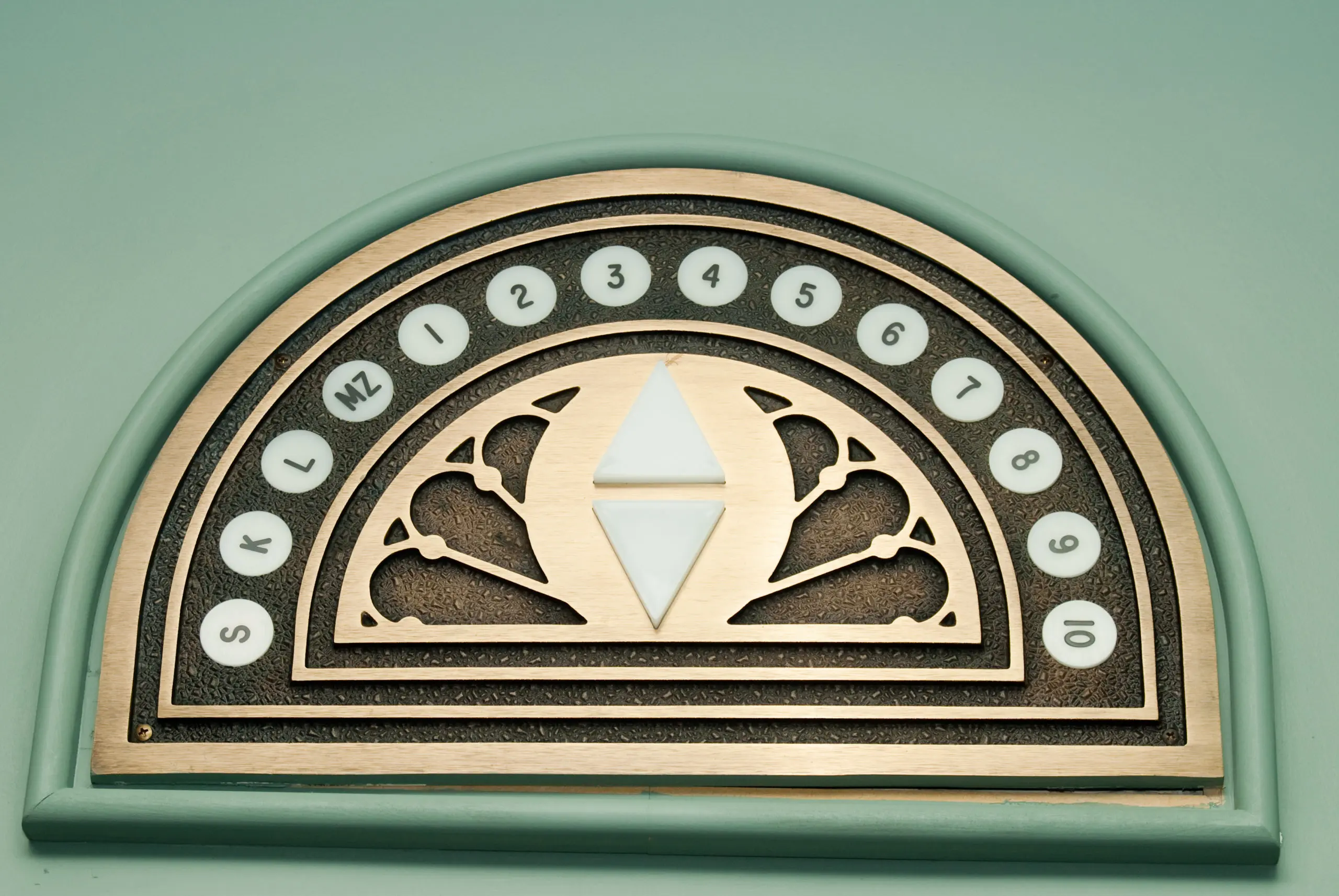 An antique semicircular brass elevator floor indicator dial mounted on a light green wall showing floor levels from 1 to 10 along with letters