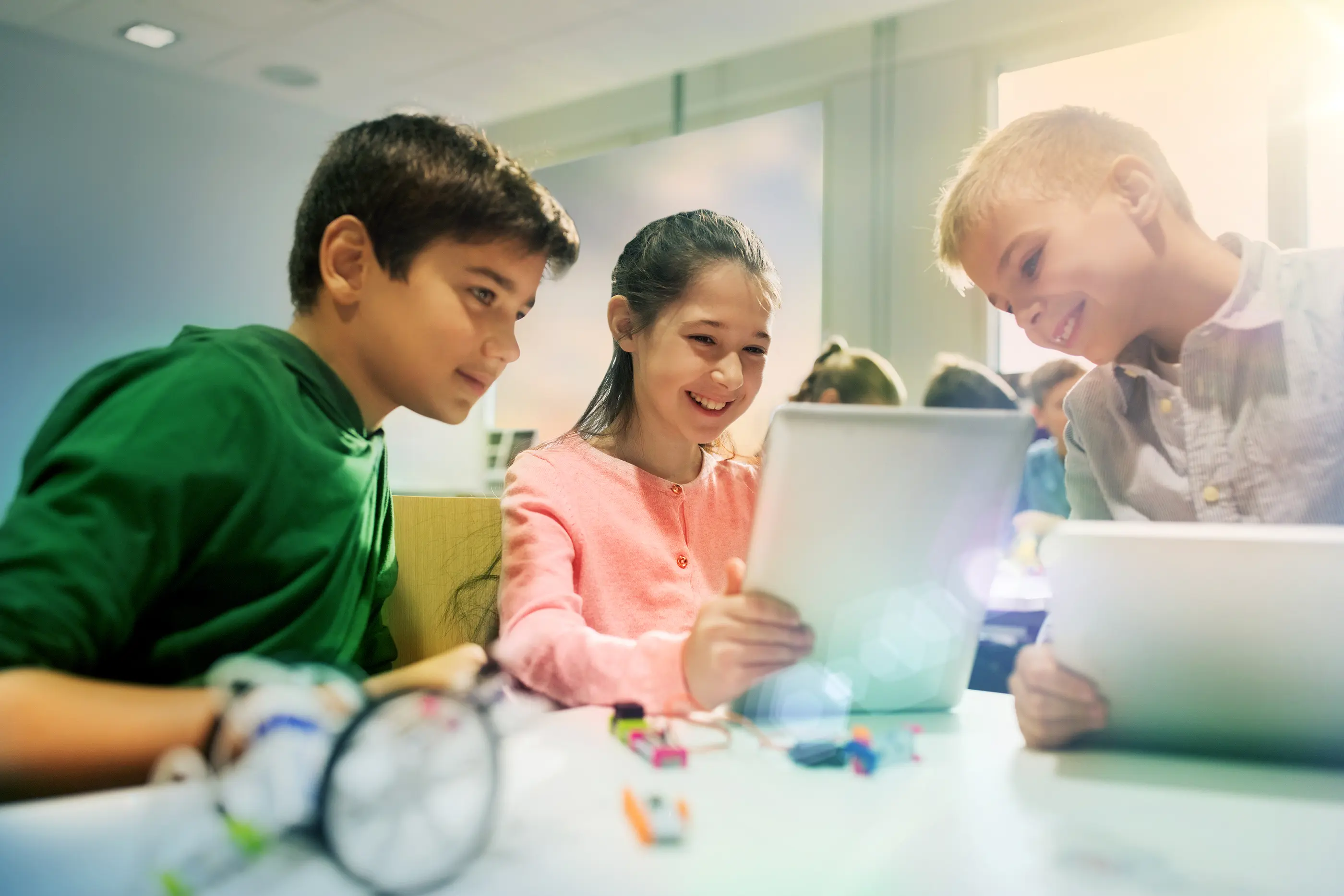 Three young students smiling and collaborating around digital tablets at a classroom table with robotics components