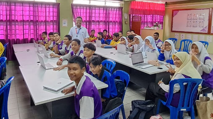 Classroom filled with kids and teacher smiling, all sitting down with laptops in front of them